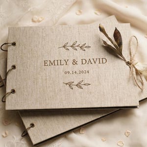 Puede incluir: Un libro de visitas cubierto de tela beige con encuadernación de anillas metálicas. La portada presenta el texto EMILY & DAVID y 14.09.2024 en tinta marrón, flanqueado por dos ilustraciones de hojas. Una rama seca está atada al lado.