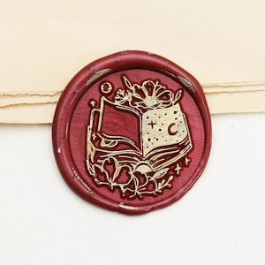 Könnte beinhalten: Ein burgunderfarbenes Wachssiegel mit einem goldgeprägten Design eines offenen Buches, Blumen, einem Mond und Sternen. Das Siegel ist kreisförmig und hat ein leichtes Vintage-Aussehen. Der Hintergrund ist hellbeige.