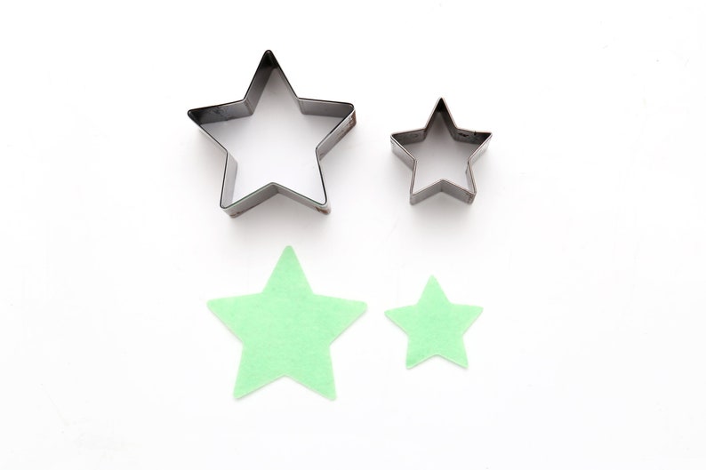 May include: Two metal star-shaped cookie cutters and two green felt stars. The larger cookie cutter is on the left, and the smaller cookie cutter is on the right. The larger felt star is below the larger cookie cutter, and the smaller felt star is below the smaller cookie cutter.