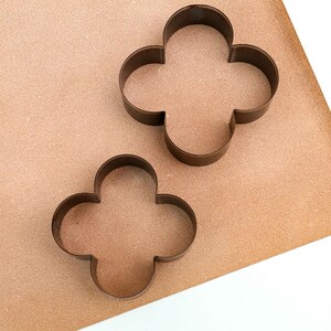 Puede incluir: Dos cortadores de galletas de metal con forma de tr&eacute;bol de cuatro hojas. Los cortadores est&aacute;n sobre una superficie marr&oacute;n texturizada.
