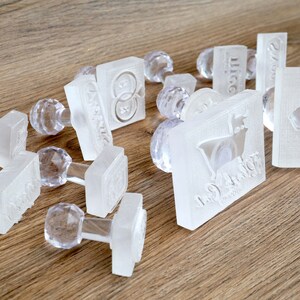May include: A collection of clear plastic stamps with various designs, including a heart, a goat, and a ring. The stamps are on a wooden surface.