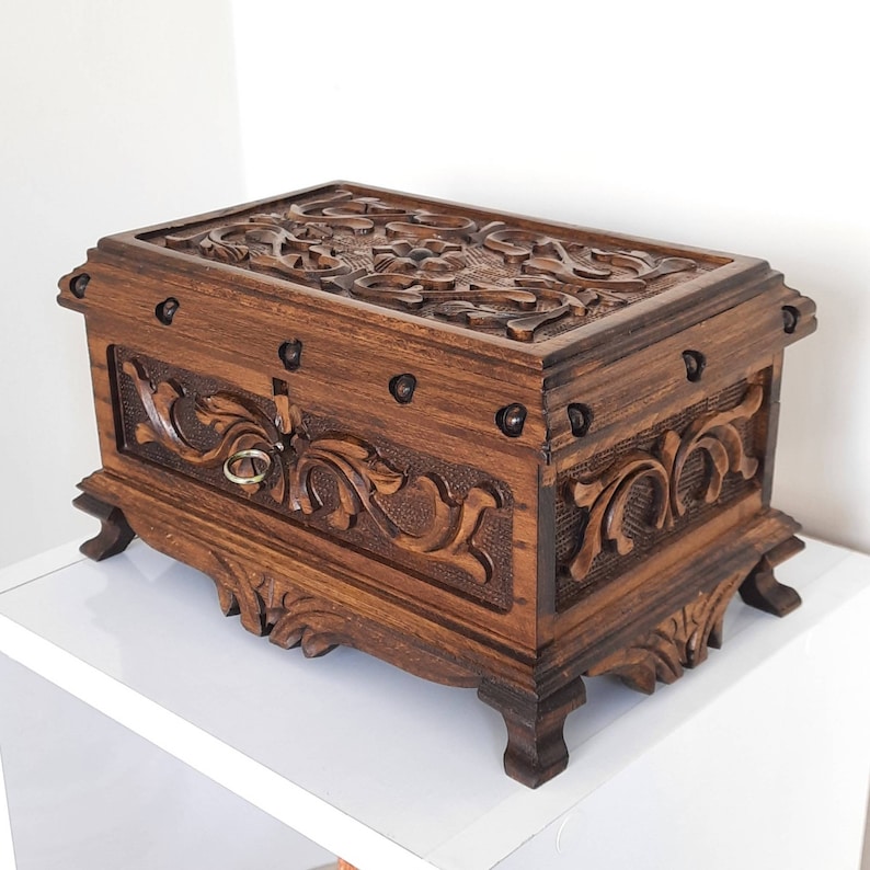 K&ouml;nnte beinhalten: Eine dunkelbraune Holzkiste mit aufwendigen Schnitzereien von floralen Mustern. Die Kiste hat eine kleine Schublade mit einem Messinggriff und ist mit Metallverzierungen verziert.