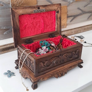 K&ouml;nnte beinhalten: Eine Holzk&auml;stchen f&uuml;r Schmuck mit aufwendigen Schnitzereien und roter Samt-Auskleidung. Die Box ist ge&ouml;ffnet und mit verschiedenen Schmuckst&uuml;cken gef&uuml;llt, darunter Halsketten, Armb&auml;nder und Ringe.