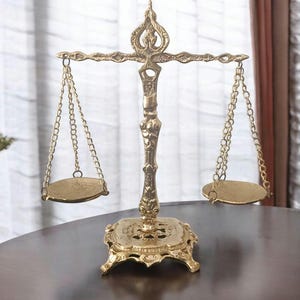 May include: A decorative brass scale with two oval pans suspended by chains. The scale has an ornate, detailed design and sits on a decorative base. The scale is a golden colour.