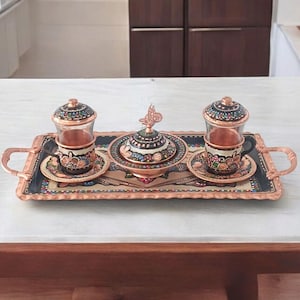 Peut inclure: Un plateau de service cuivré avec deux tasses à thé, des soucoupes et un sucrier. Les tasses et le sucrier présentent des motifs floraux complexes. Le plateau a des poignées et une bordure décorative. L'ensemble est posé sur une surface blanche.