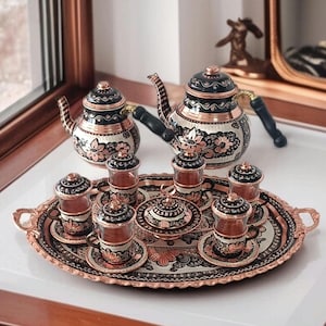 Puede incluir: Un juego de té de cobre ornamentado con detalles en negro y plateado. El juego incluye dos teteras, seis vasos pequeños, platillos y un azucarero cubierto, todo dispuesto sobre una bandeja ovalada. El juego tiene un diseño floral.
