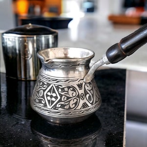 Può includere: Una caffettiera turca d'argento con un intricato disegno floreale. La caffettiera ha un manico in legno e si trova su una superficie nera.