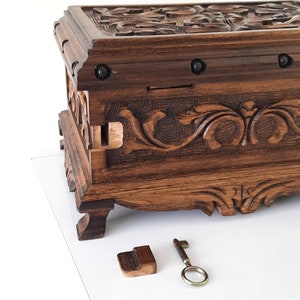 K&ouml;nnte beinhalten: Eine Holzkiste mit aufwendigen Schnitzereien und einem Schl&uuml;ssel. Die Kiste hat eine dunkelbraune Oberfl&auml;che und ist mit floralen Mustern verziert. Der Schl&uuml;ssel ist aus Metall und hat ein schlichtes Design.