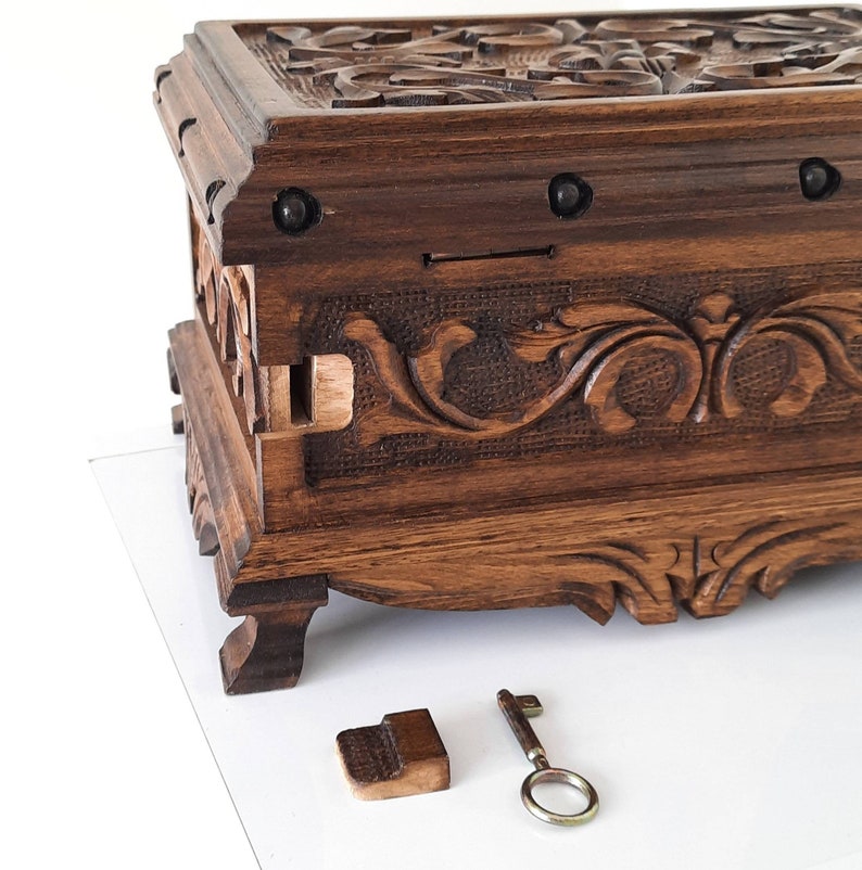 K&ouml;nnte beinhalten: Eine Holzkiste mit aufwendigen Schnitzereien und einem Schl&uuml;ssel. Die Kiste hat eine dunkelbraune Oberfl&auml;che und ist mit floralen Mustern verziert. Der Schl&uuml;ssel ist aus Metall und hat ein schlichtes Design.