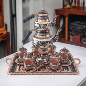 May include: A copper tea set with a double-tiered teapot, cups, saucers, and a serving tray. The set features intricate floral and geometric patterns in black, white, and blue. The teapot has black handles, and the cups have glass sides.