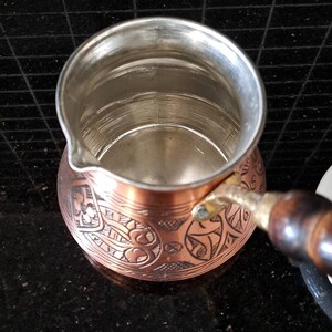 May include: A copper Turkish coffee pot with an ornate engraved design. The pot has a wooden handle and a shiny interior.