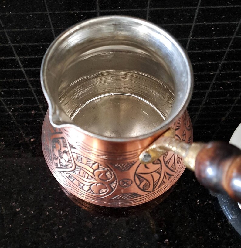 May include: A copper Turkish coffee pot with an ornate engraved design. The pot has a wooden handle and a shiny interior.