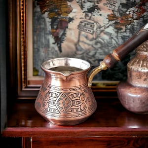 May include: A traditional copper coffee pot with an ornate design and a wooden handle. The pot is filled with coffee and is sitting on a wooden surface.