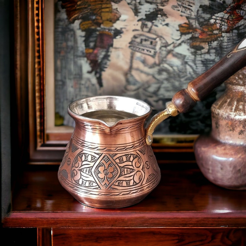 May include: A traditional copper coffee pot with an ornate design and a wooden handle. The pot is filled with coffee and is sitting on a wooden surface.