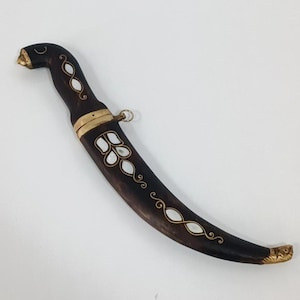 May include: A decorative dagger with a curved blade and dark brown handle. The handle is adorned with white inlaid designs and gold accents. The sheath is dark brown with gold trim and a small ring. The tip of the blade has gold detailing.