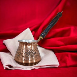 Könnte beinhalten: Eine Kaffeekanne aus Kupfer mit einem verzierten Design und einem Holzgriff steht auf einem weißen Tuch vor einem roten Hintergrund.