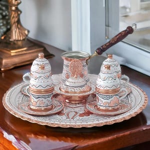 May include: A white and copper Turkish coffee set with intricate floral designs. The set includes a coffee pot, two cups, two saucers, and a sugar bowl. The set is on a round tray with a scalloped edge.