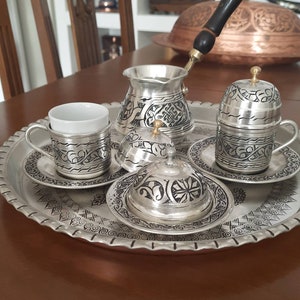 May include: A silver coffee set with intricate etched designs, including a coffee pot, cups, saucers, and covered dishes, arranged on a decorative tray. The set features a dark handle on the coffee pot and small gold-colored accents.
