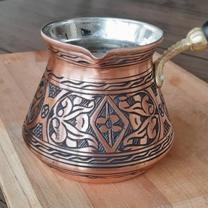 May include: A traditional copper Turkish coffee pot with a black floral design and a wooden handle. The pot is on a wooden surface.