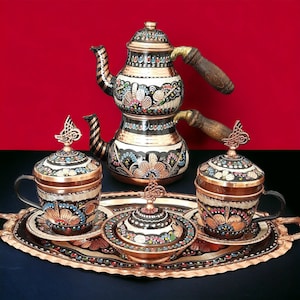 Könnte beinhalten: Ein traditionelles türkisches Kaffee-Set mit einer Kupferkanne und zwei Tassen mit Deckeln. Die Kanne und die Tassen sind mit bunten Blumenmustern verziert. Das Set wird auf einem Kupfertablett mit einem dekorativen Rand präsentiert.
