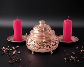 May include: A copper-colored decorative container with a lid, intricate etched designs, and small feet. Two red pillar candles stand on matching copper saucers, creating a warm ambiance. The arrangement is set against a dark background.