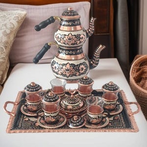 May include: A copper tea set with a double-tiered teapot and a serving tray. The set includes small glasses, saucers, and covered cups. The teapot and tray feature black and floral designs, with black handles and accents.