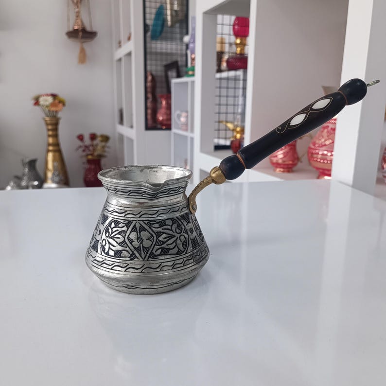 May include: A silver Turkish coffee pot with a black wooden handle and intricate floral designs. The pot has a brass spout and a rounded body.