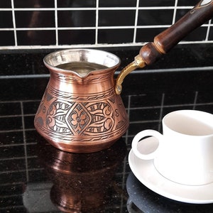 May include: A copper coffee pot with an ornate design and a wooden handle. The pot is filled with water and is sitting on a black countertop. A white coffee cup and saucer are in the foreground.