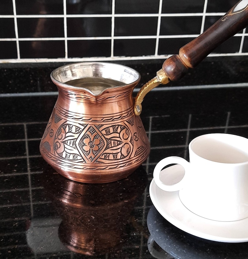 May include: A copper coffee pot with an ornate design and a wooden handle. The pot is filled with water and is sitting on a black countertop. A white coffee cup and saucer are in the foreground.