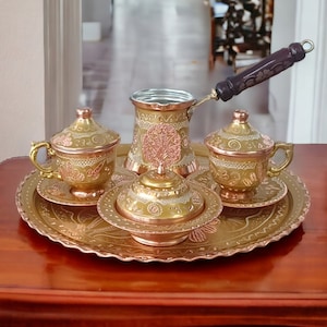 Puede incluir: Un juego de café turco tradicional con una olla de cobre, dos tazas, un azucarero y una bandeja. El juego está decorado con intrincados patrones florales y tiene un acabado dorado.