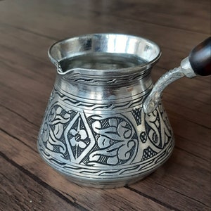 May include: A silver Turkish coffee pot with an ornate floral design. The pot has a wooden handle and is filled with water.