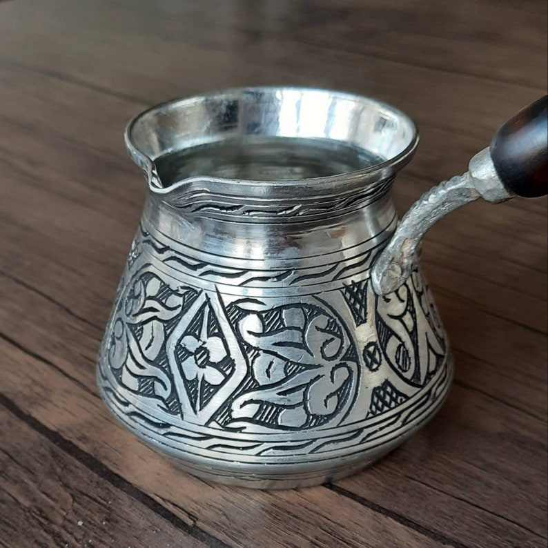 May include: A silver Turkish coffee pot with an ornate floral design. The pot has a wooden handle and is filled with water.