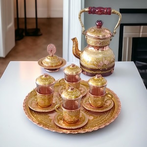 May include: A set of copper teacups and saucers with intricate gold designs, a matching teapot with a wooden handle, and a sugar bowl with a lid. The set is arranged on a decorative tray with a gold and copper finish.