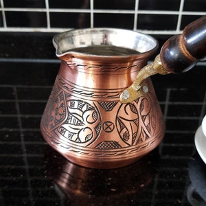 May include: A copper coffee pot with a wooden handle and intricate floral designs. The pot has a silver lining and is sitting on a black tiled surface.