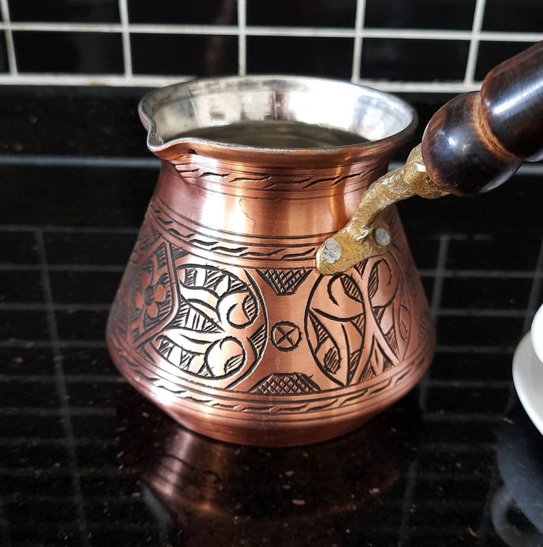 May include: A copper coffee pot with a wooden handle and intricate floral designs. The pot has a silver lining and is sitting on a black tiled surface.