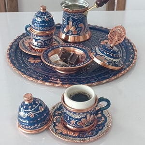 Puede incluir: Un juego de café turco tradicional con un diseño de cobre y azul. El juego incluye una cafetera, dos tazas y platillos, un azucarero y una bandeja. Las tazas y platillos tienen un interior blanco y un exterior azul y cobre. La bandeja es azul con un borde de cobre.