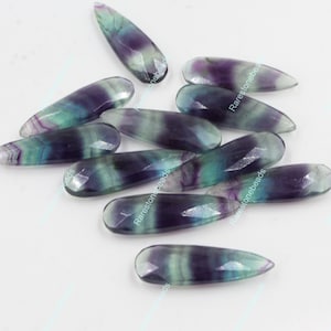 May include: A group of ten teardrop-shaped fluorite beads in shades of green, purple, and white. The beads are faceted and have a glossy finish.