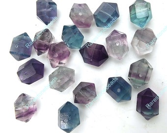 May include: A collection of faceted fluorite beads in various colors, including purple, blue, green, and clear. Each bead has a hexagonal shape with a drilled hole for stringing. The beads are scattered across a white surface.