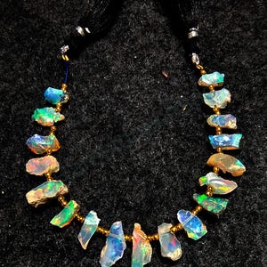 Könnte beinhalten: Eine Halskette aus schillernden Opal-Steinen, die auf einer goldenen Kette mit goldenen Perlen aufgefädelt sind. Die Halskette hat an jedem Ende schwarze Quasten.