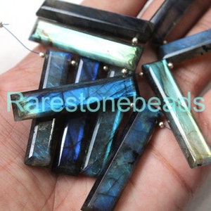 Op de afbeelding: Een verzameling van iriserende blauwe en groene labradorit edelsteen kralen, elk gesneden in een rechthoekige vorm. De kralen zijn in een stapel gerangschikt, waardoor hun unieke schittering en glans zichtbaar worden.