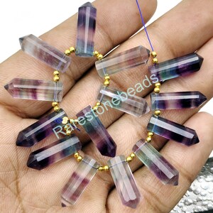 Op de afbeelding: Een streng facetgeslepen fluorite edelsteen kralen in tinten van paars, groen en blauw. De kralen zijn geregen op een gouden draad met kleine gouden afstandhouders. De kralen zijn allemaal ongeveer 2,5 cm lang en hebben een puntige punt.