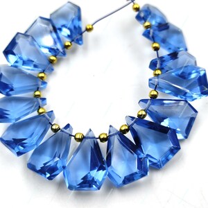 May include: A strand of blue gemstone beads with a faceted, teardrop shape. The beads are strung on a thin, clear elastic cord with gold-colored spacers between each bead.