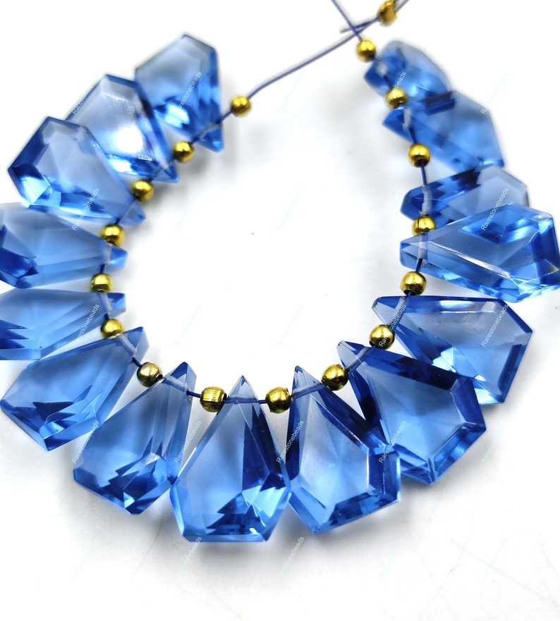 May include: A strand of blue gemstone beads with a faceted, teardrop shape. The beads are strung on a thin, clear elastic cord with gold-colored spacers between each bead.
