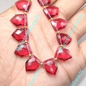 May include: A strand of faceted, deep red gemstone beads. Each bead is a geometric shape with multiple facets, strung together with gold-colored spacers. The beads are displayed against a light background.