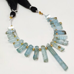 May include: A necklace featuring light blue aquamarine gemstone beads. The beads are cylindrical and strung together with gold-colored spacers. The necklace is displayed on a white surface, with black string and tassels at the top.
