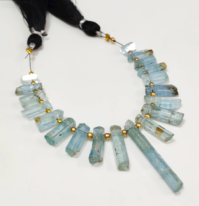 May include: A necklace featuring light blue aquamarine gemstone beads. The beads are cylindrical and strung together with gold-colored spacers. The necklace is displayed on a white surface, with black string and tassels at the top.