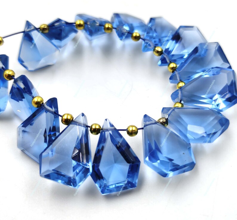 May include: A strand of blue gemstone beads with gold accents. The beads are faceted and have a teardrop shape. The beads are strung on a thin wire.