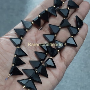 Puede incluir: Una hebra de cuentas de piedra preciosa negra en forma de triángulo con acentos dorados. Las cuentas están enhebradas en un alambre y tienen aproximadamente 1 cm de tamaño. Las cuentas se muestran en una mano.