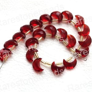 Pode incluir: Um fio de contas de cristal vermelho em forma de luas crescentes. As contas são enfiadas em um fio de ouro.