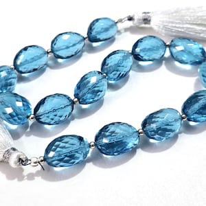 May include: Strand of faceted, oval-shaped blue gemstones. Each gem is connected by a small silver bead. White tassels are attached to each end of the strand. The gems have a transparent, glassy appearance.